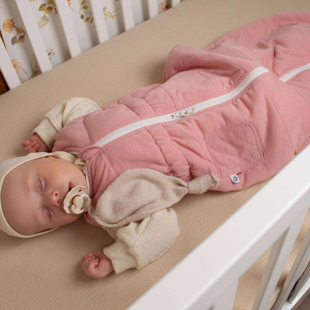 Baby sover i en rosa Easygrow nattpose over en overmadrass i ull. Under nattposen er baby kledd i en langermet, kremhvit ullpysj og en kremhvit tynn babylue. Baby sover med smokk i munn. Den har en strikket koseklut hengene ned fra håndtaket. Baby sover med armene opp