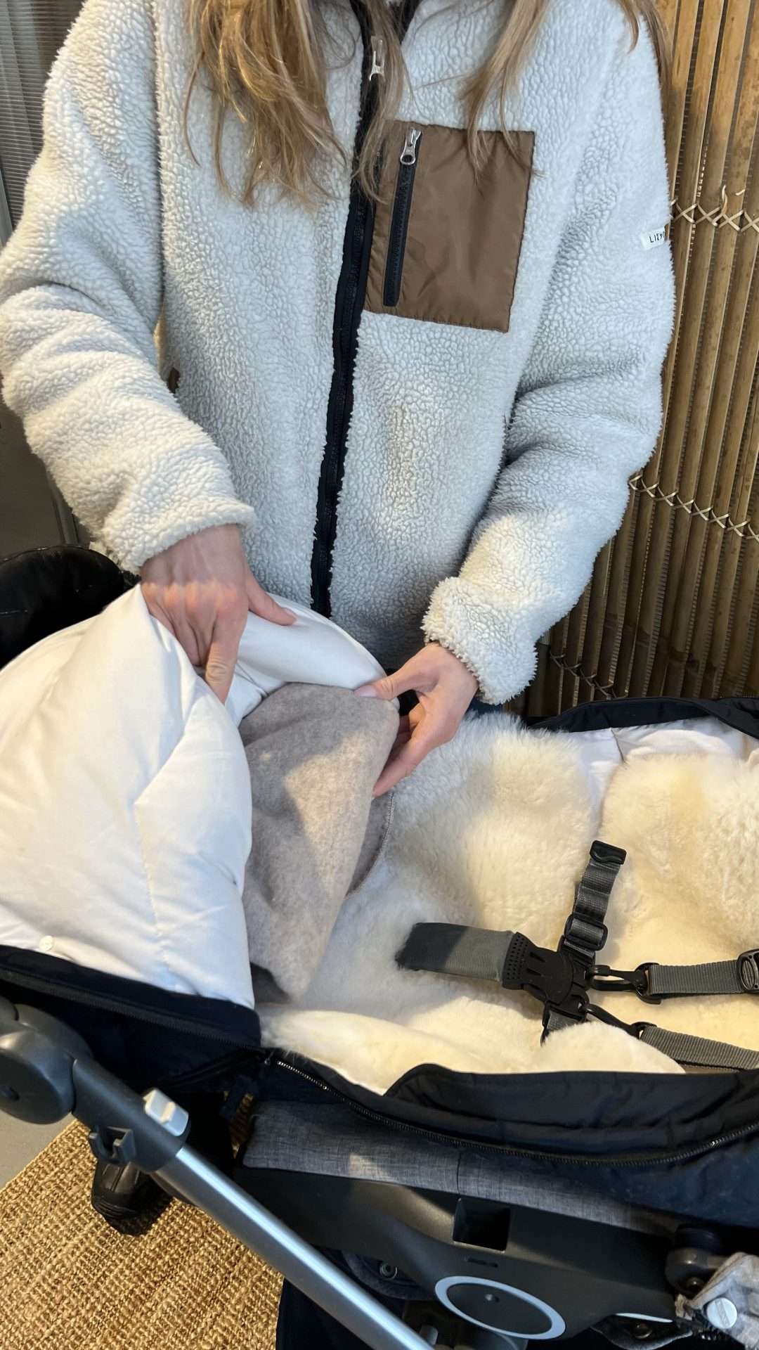 Dame fyller en vognpose med ullpledd i fotenden. Vognposen har også et lammeskinn på innsiden av seg. Stroppene på barnevognen er tredd igjennom lammeskinn og vognposen.