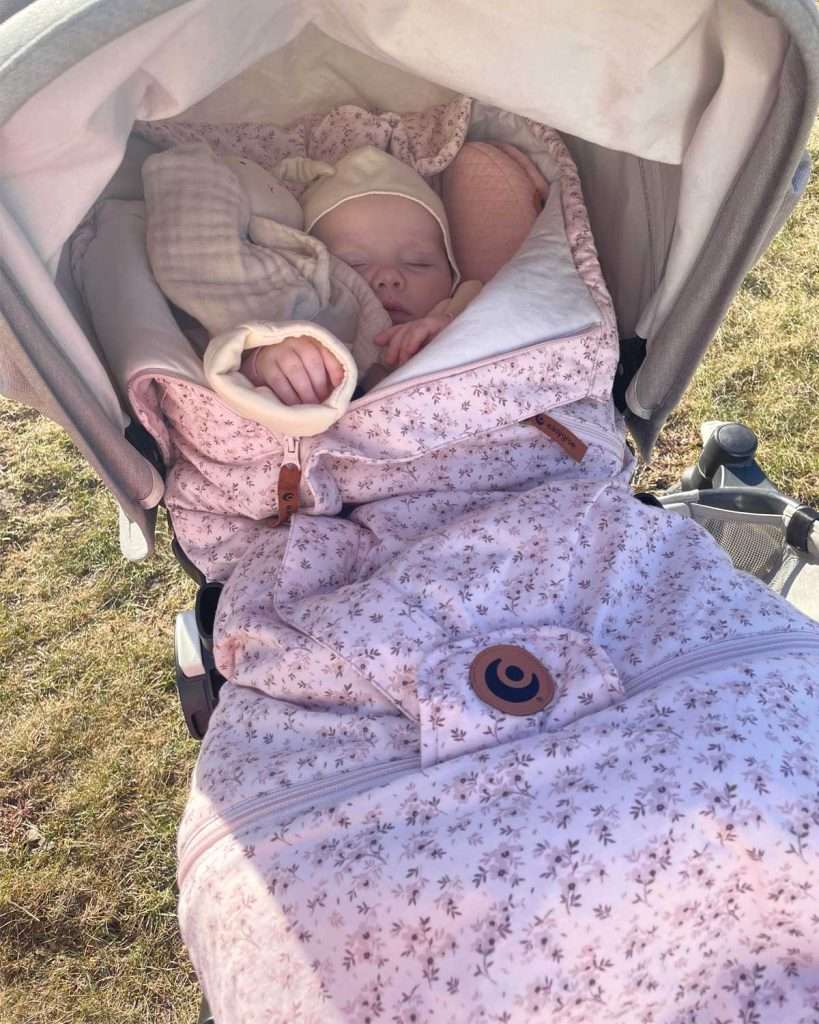 Baby sover ute i vogn pakket i en rosa Easygrow Vognpose med en Easygrow Minimizer under seg. Minimizer er puttet på innsiden av vognposen, ikke under den. Barnet er kledd i kremhvit klær og har en koseklut med bamse ved siden av seg i samme kremhvit farge. Det ser ut som at det er vår ute. Solen skinne og gresset begynner er grønt. Men barnet er kledd litt for godt til at det er en varm sommerdag.