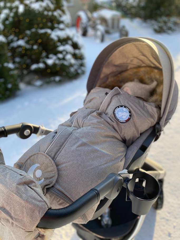 Grå barnevogn med grå Easygrow Nature vognpose oppi og grå Easygrow Håndmuffer festet til håndtaket står ute på vinteren. Baby sover i vognen, litt av ansiktet og en beige ullue med ører synes opp fra vognposen. Vognen står ute på en vei med masse snø.