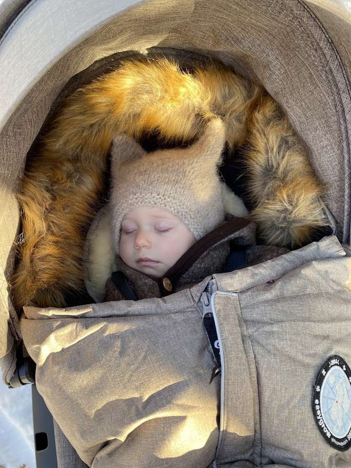 Baby sover i en beige vognpose av typen Easygrow Nature. Baby er kledd i en beige balaclava med ører strikket i ull og en brun ulldress. Under baby ser vi et lammeskinn som er plassert oppi vognposen. Easygrow nature vognpose har fuskepels rundt hetten.