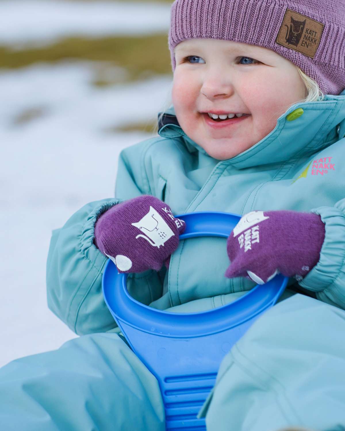 Lite barn ute og aker i snøen kledd i Kattnakkeen yttertøy. Blå vinterdress, lilla ullue og lilla magiske ullvotter.