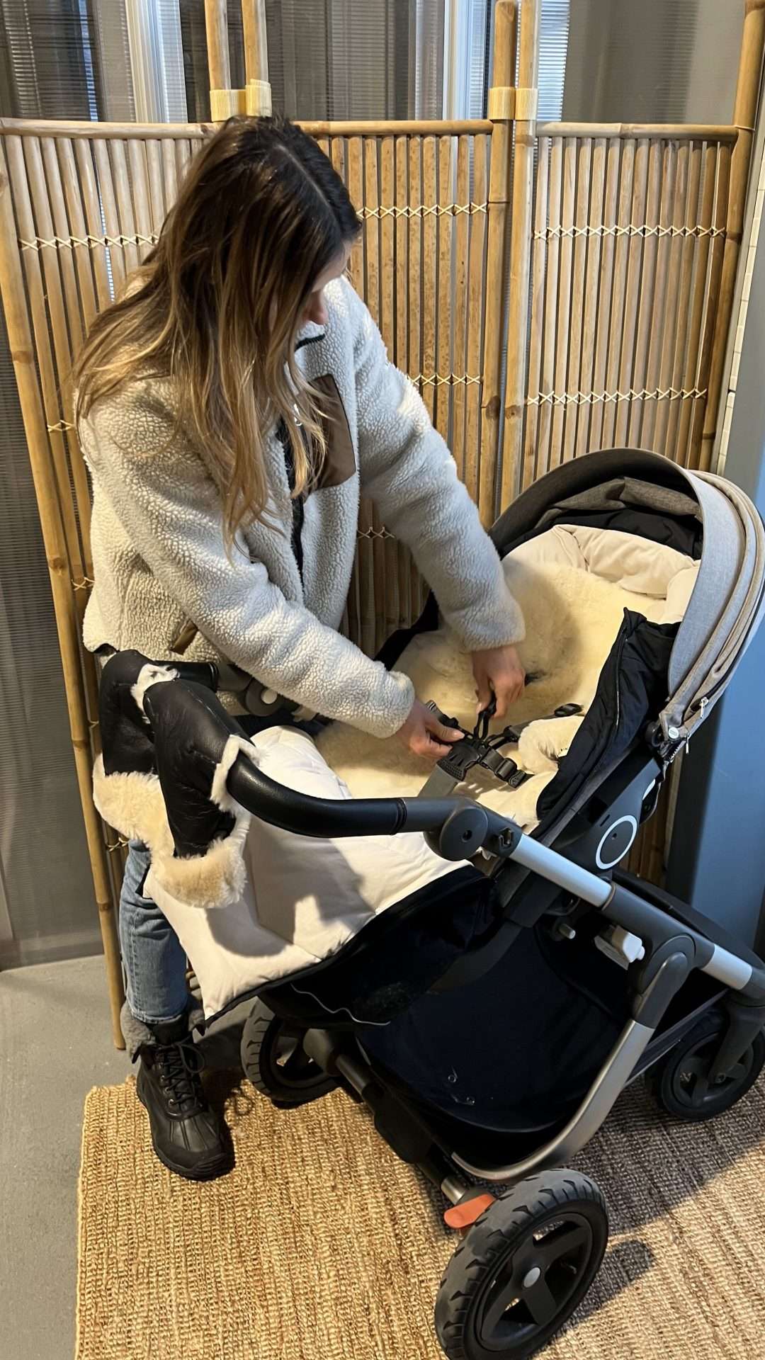 dame står og monterer hvitt lammeskinn til vogn på innsiden av en svart voksi vognpose. Barnevognen er grå. Vognen står inne i en gang med bambus vegg.