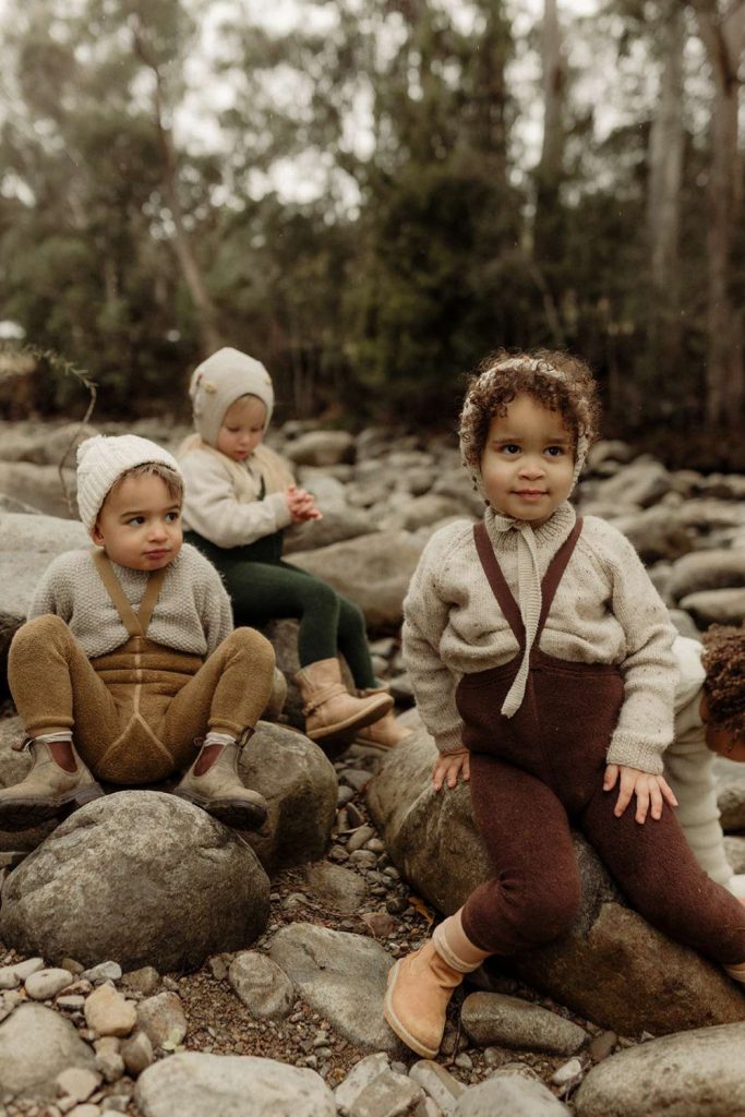 Tre barn sitter ute på steiner. Alle tre har på seg en lik strømpebukse med seler over en grå ullgenser. Strømpebukser er i ulike farger. En brun, en beige og en grønn.
