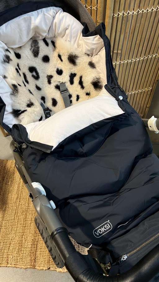 Svart Voksi Vognpose Urban med et lammeskinn til vogn i dalmatiner mønster plassert på innsiden. Vognposen er montert oppi en grå barnevogn som står inne i en gang.