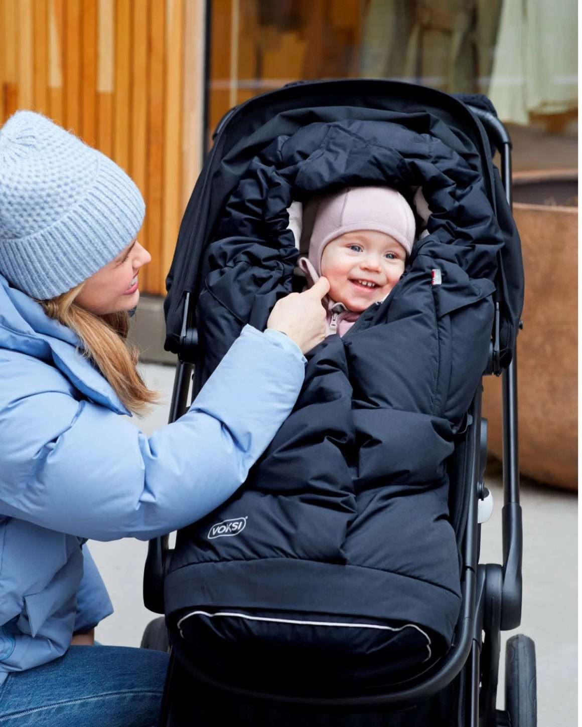 Lite barn oppi en svart Voksi City North vinterpose. Barnet sitter ute i en sittedel på en sporty barnevogn.
