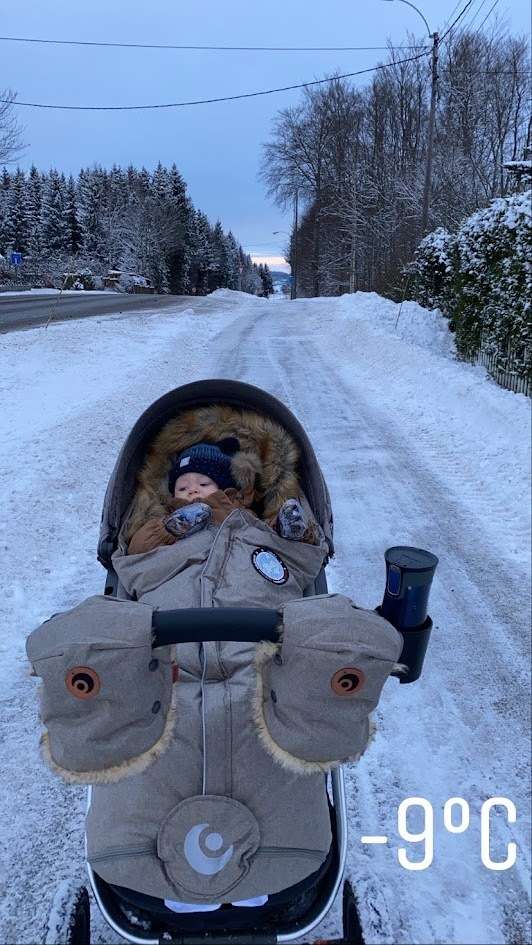 Baby er ute i minus 9 grader, det står det på bilde. Baby sitter i en barnevogn med Easygrow Natur vognpose, en brun Vinterdress og en blå balaclava lue. På håndtaket av vognen er det festet Easygrow Håndmuffer til den som skal trille.