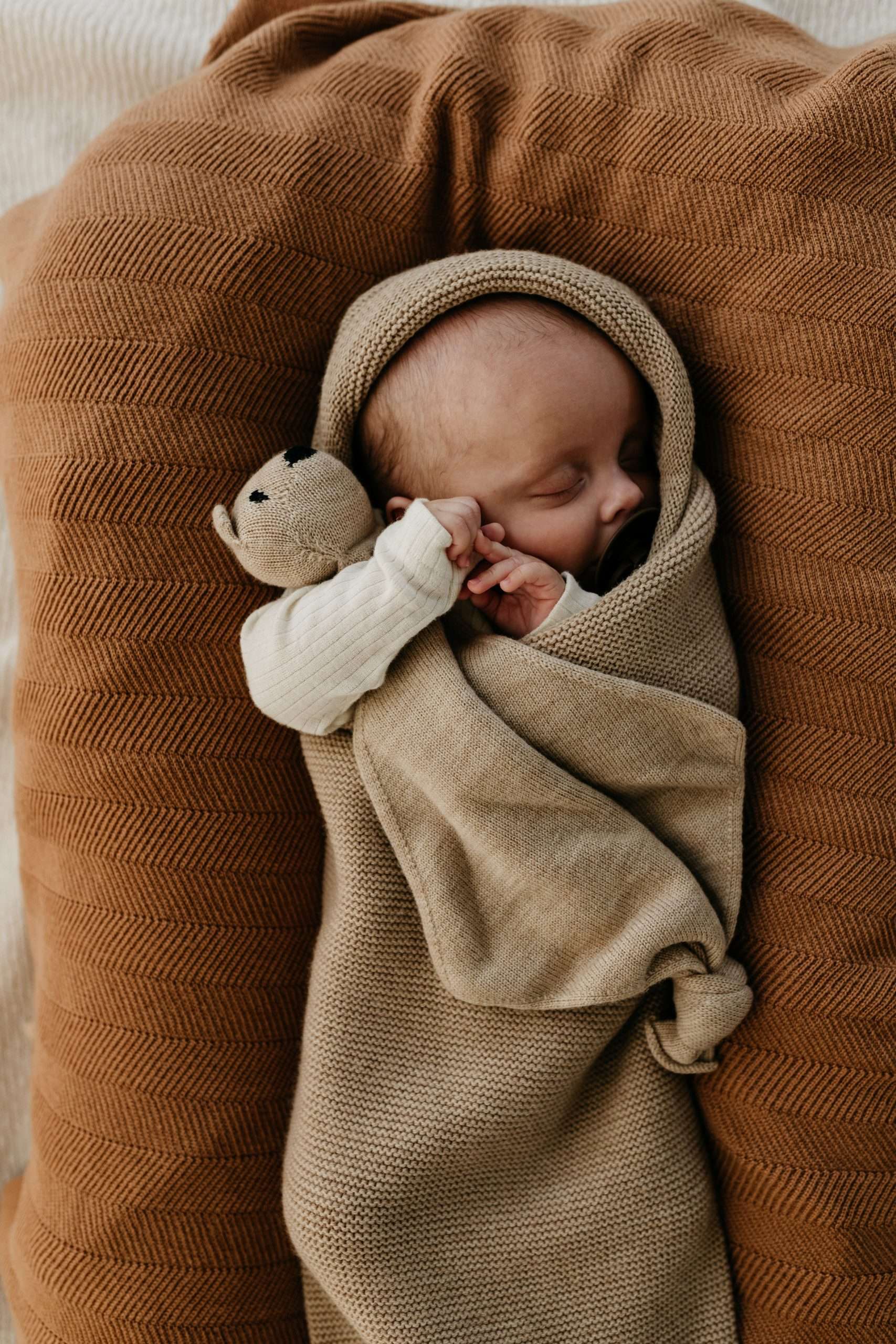 Baby sover i et babynest pakket inn i en beige svøpepose i ull og koser på en koseklut med bamse av samme ullgarn som svøpeposen.