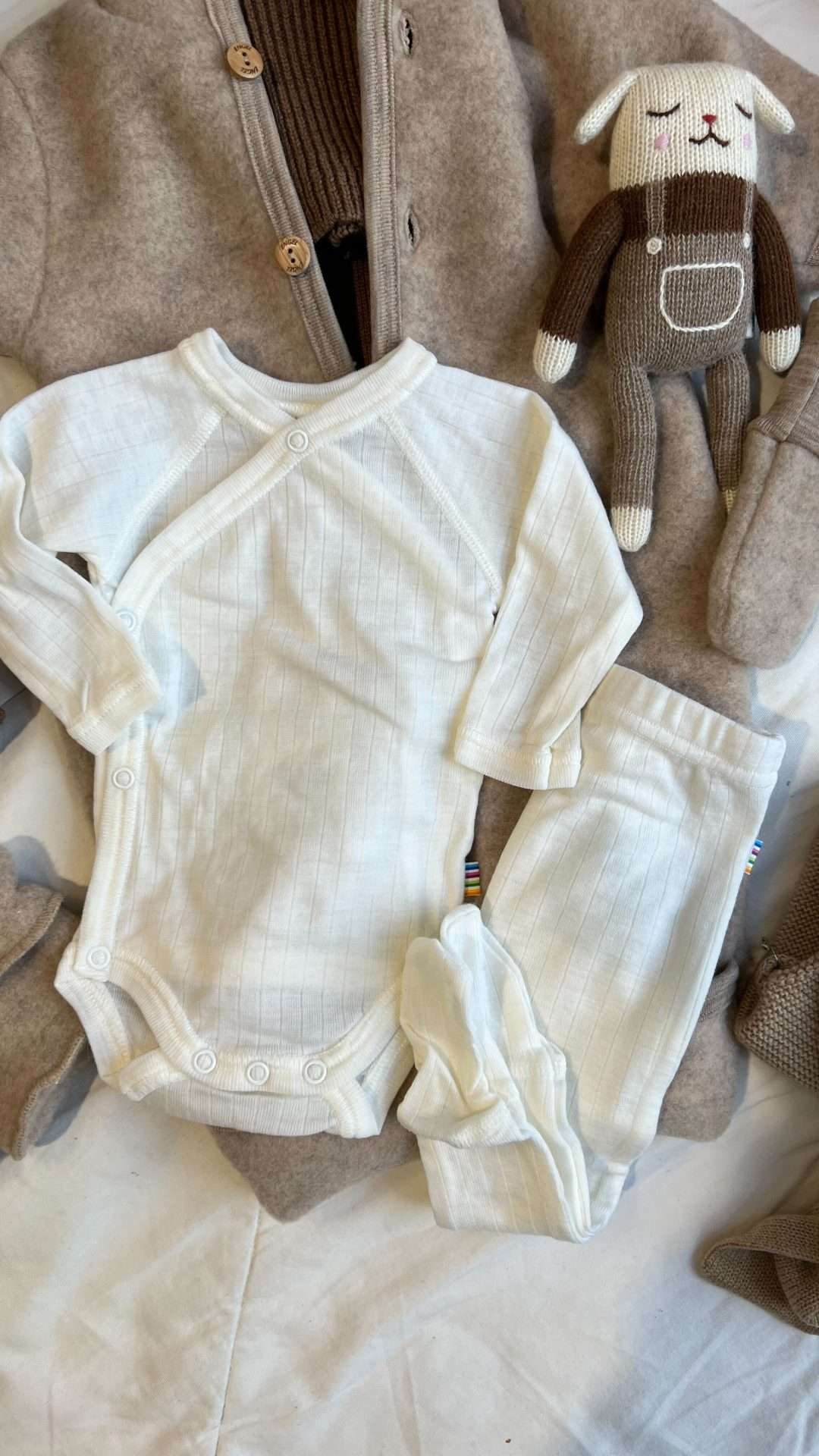 Hvite ullklær til nyfødt. Omslagsbody og ulltights med fot. Ved siden av ligger et lite kosedyr som er strikket i ull. kosedyret er et lite lam med brun genser og beige snekkerbukse.