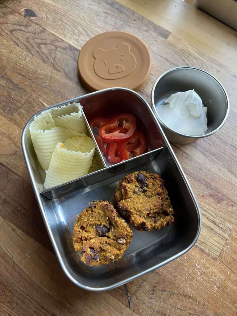 Metaøll matboks med flere rom fylt med to matmuffins, gulostskiver og paprika. Ved siden av står en liten, rund matboks fylt med rømme.