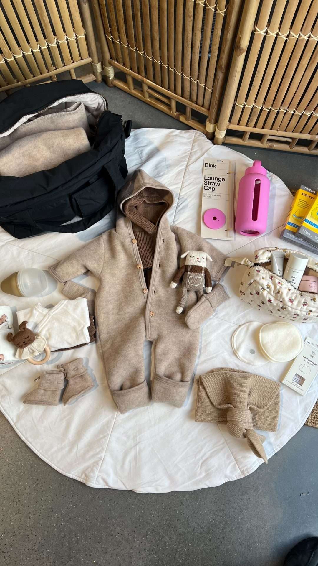 Tips til fødebaginnhold av klær til nyfødt og ting til mamma. Til babymamma er det vannflaske, toalettmappe, ammeinnlegg, amme bh og klær. Til baby et hentesett i ull, bamse, ullpose, ulltøfler og et par omslagsbody i ull. Tingene ligger spredt utover en kremhvitt teppe. Ved siden av står en svart stelleveske som er klar til å bli fylt med dette innholdet