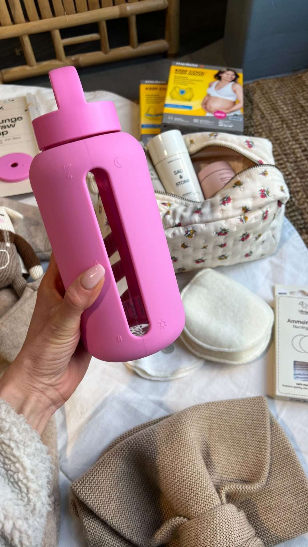 Rosa drikkeflaske med chunky lokk med håndtak. I bakgrunnen synes annet fødebag innhold; toalettmappe, ammeinnlegg, amme bh og ullklær til nyfødt.
