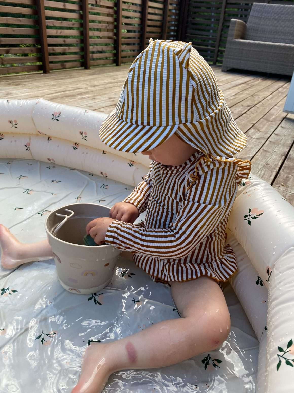 Lite barn sitter i oppblåsbart badebasseng med uv drakt og badehatt fra Liewood på seg. Uv drakt er av type badedrakt med lange armer. Dette er et matchende sett i brune og hvite striper.